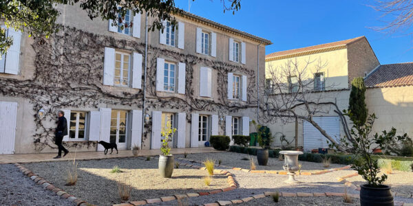 C-IMG_0348-sm Traditional stone house with white shutters and ivy-covered facade, person walking with black dog across gravel courtyard with decorative plantings under clear blue sky. Credit: James Judd