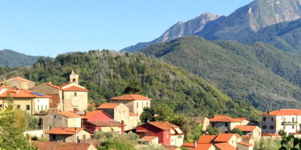 Marianne-Ryan-art-course-in-Italy publ image terenzano-1 copy Italian hillside village with terracotta roofs and cream-colored buildings nestled among lush green hills, with a dramatic rocky mountain peak rising in the background under blue sky. Credit: Marianne Ryan